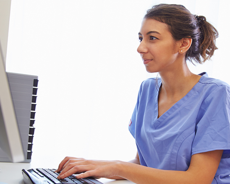 A nurse sitting by a desk in front of a computer while typing in the keyboard A nurse sitting by a desk in front of a computer while typing in the keyboard